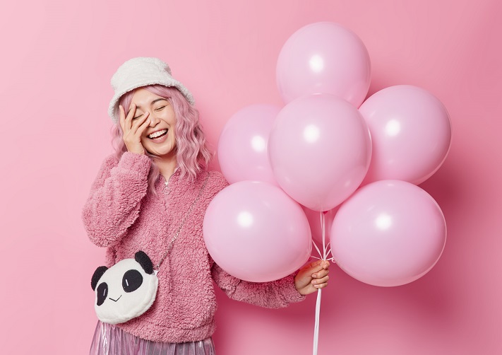 positive woman with pink hair keeps hand face smiles gladfully dressed outerwear has fun special occasion enjoys celebration holds bunch inflated balloons isolated pink wall